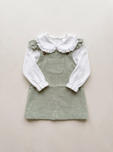 Load image into Gallery viewer, Close flat-lay view of a sage green corduroy pinafore dress and white blouse, showing the ruffle straps, front pocket and lace-trimmed collar.