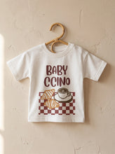 Load image into Gallery viewer, Organic cotton kids t-shirt with Babyccino coffee and croissant illustration hanging on wooden hanger against neutral wall