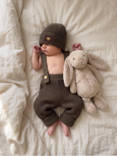 Load image into Gallery viewer, Newborn wearing oversized khaki knit braces trousers and a matching knit beanie, lying on an ivory linen bed next to a beige plush bunny.