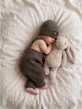 Load image into Gallery viewer, Baby wearing khaki knit braces trousers with rolled cuffs and a matching khaki knit beanie, sleeping beside a beige plush bunny on a soft ivory linen bed.