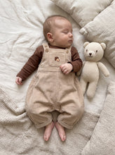 Load image into Gallery viewer,     Sleeping baby wearing beige corduroy acorn dungarees with a brown long sleeve top, lying beside a knitted teddy bear.
