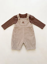 Load image into Gallery viewer,     Beige corduroy baby dungarees with acorn embroidery layered over a brown long sleeve top, flat lay on an off-white background.
