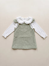 Load image into Gallery viewer, Sage green corduroy pinafore dress with a front pocket and wooden-look buttons, styled with a white long-sleeve blouse with a lace-trimmed collar.
