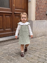 Load image into Gallery viewer, Toddler wearing a sage green corduroy pinafore dress with a white lace-collar blouse, standing in front of a wooden doorway.

