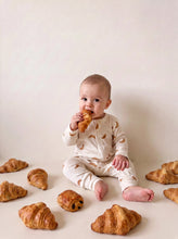 Load image into Gallery viewer, Baby wearing the Croissant organic cotton sleepsuit while holding a croissant
