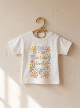 Load image into Gallery viewer, Organic cotton kids t-shirt with colourful floral Isn’t She Lovely graphic hanging on wooden hanger against neutral wall
