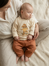 Load image into Gallery viewer, Baby wearing organic cotton silly goose graphic top with ribbed rust trousers while sitting on bed
